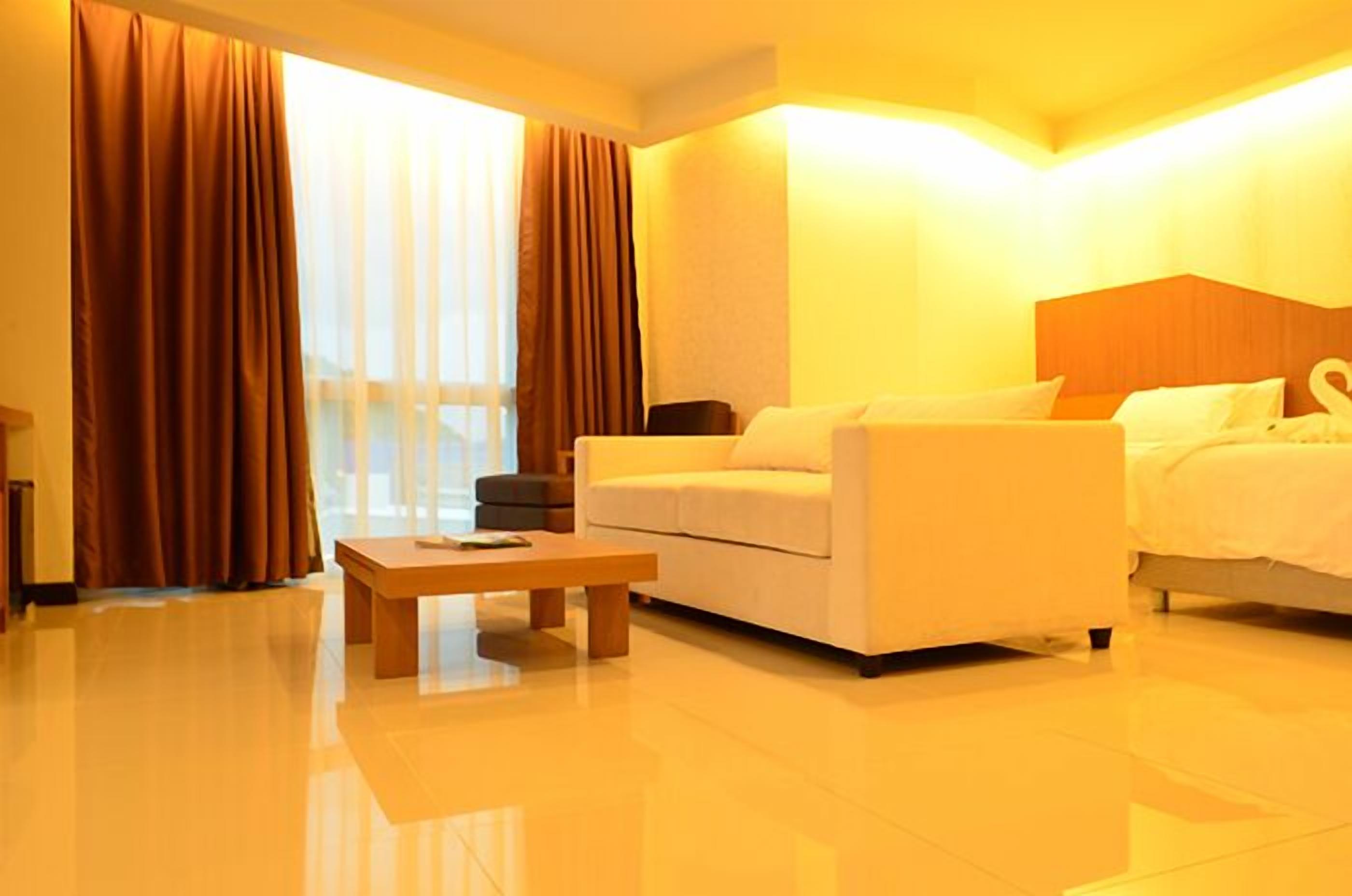 A hotel room with a sofa, a bed, a wooden coffee table, and a large window with curtains.