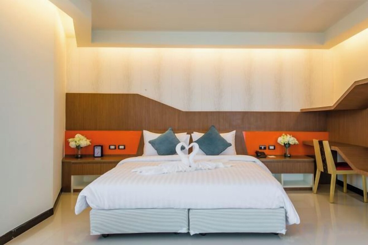 Een moderne hotelkamer met een groot bed, een houten hoofdbord, een nachtkastje met een bloevas en een bureau met een stoel.