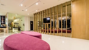 A modern hotel lobby with a reception desk, a television mounted on a wooden panel, and a seating area with red upholstered benches.