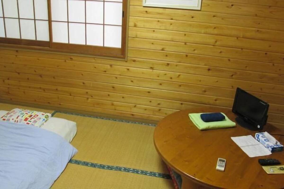 A room with wooden walls and tatami flooring, a bed with a blue blanket, a round wooden table with a TV, and a window with a grid pattern.