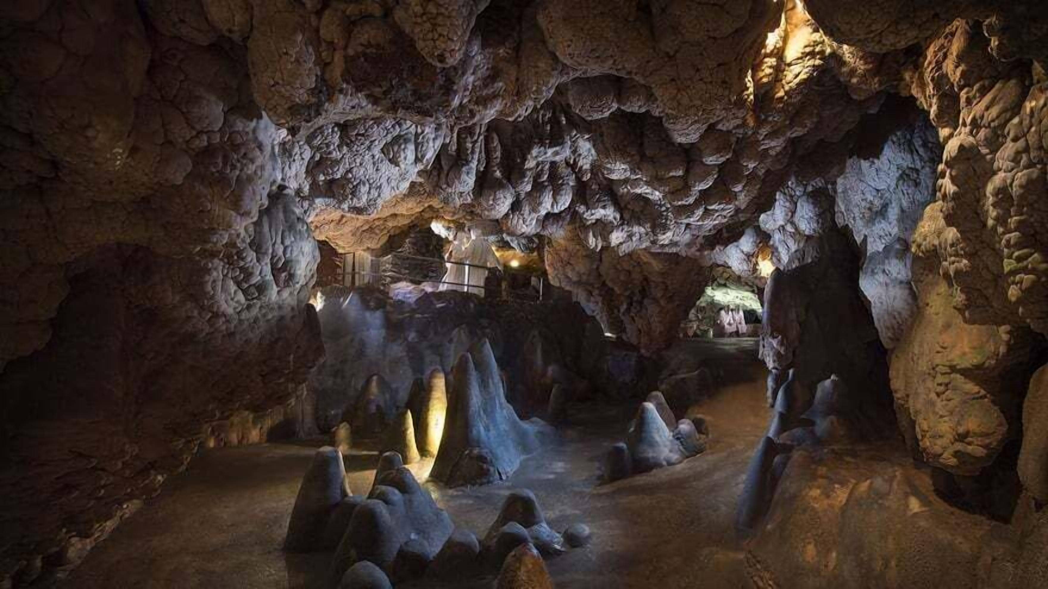 Una cueva con estalactitas y estalagmitas, iluminación tenue y un sendero que atraviesa la cueva.