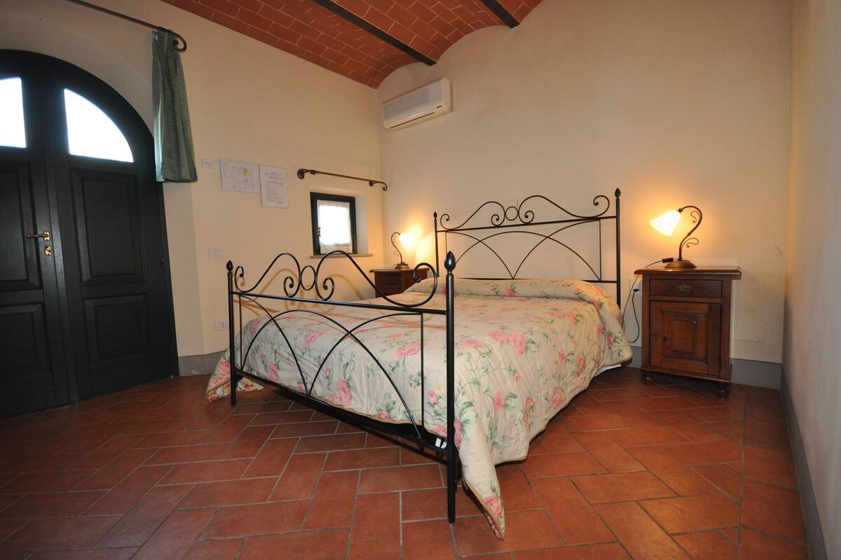 A bedroom with a metal bed frame, floral bedding, a bedside table with a lamp, and a small cabinet.