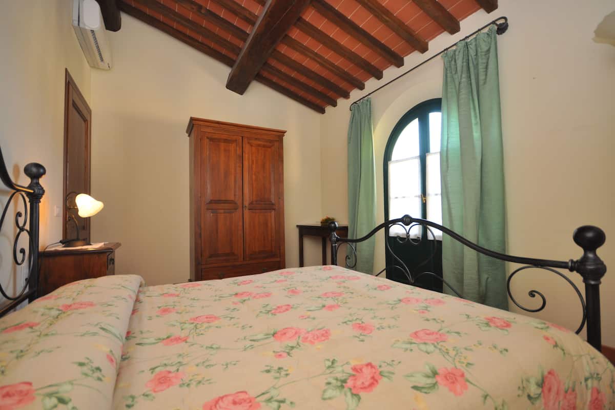 A bedroom with a floral bedspread, a wooden wardrobe, a nightstand with a lamp, and a window with green curtains.