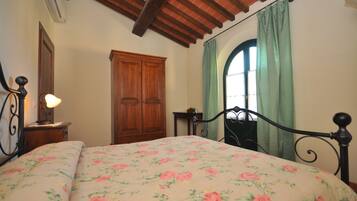 A bedroom with a floral bedspread, a wooden wardrobe, a nightstand with a lamp, and a window with green curtains.