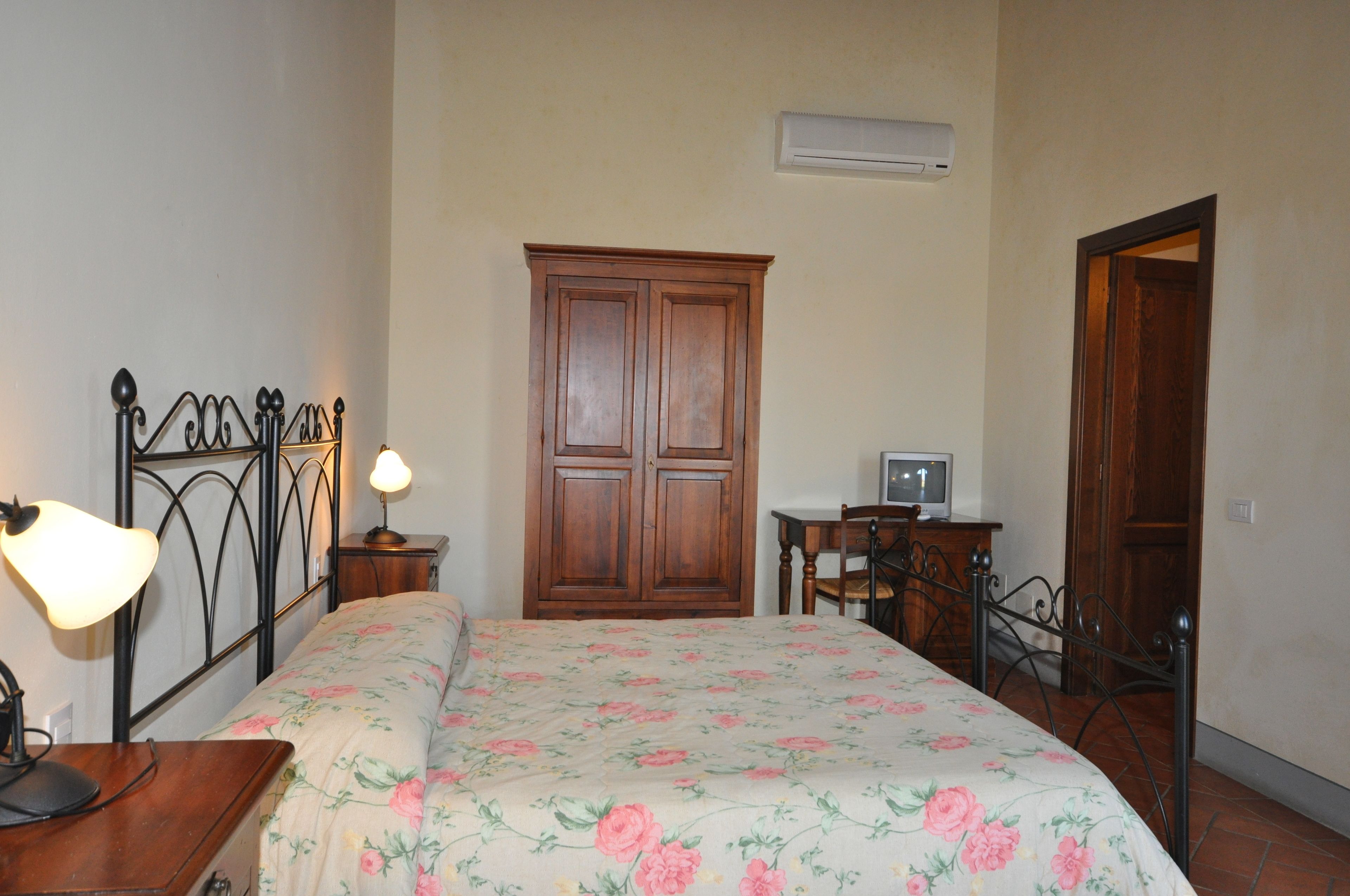A bedroom with a bed, a wooden wardrobe, a desk with a chair, a lamp, and a television.