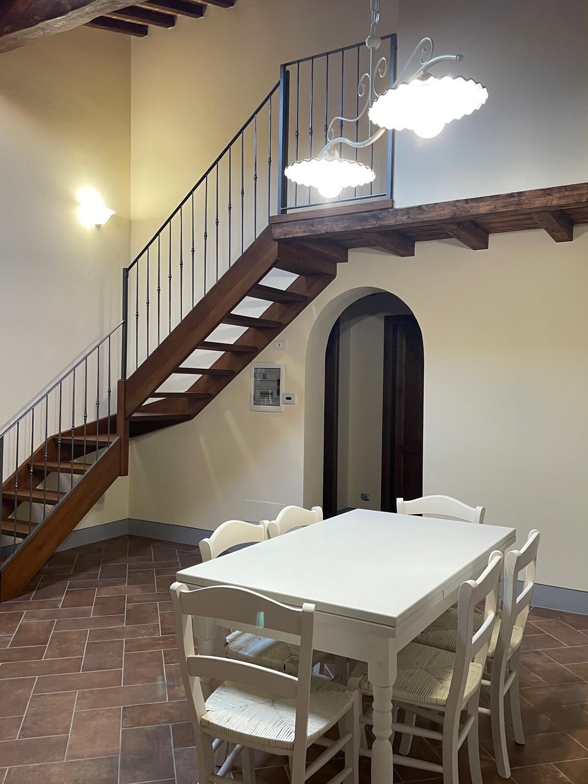 A dining area with a white table and chairs, a wooden staircase with a metal railing, and a ceiling light fixture.