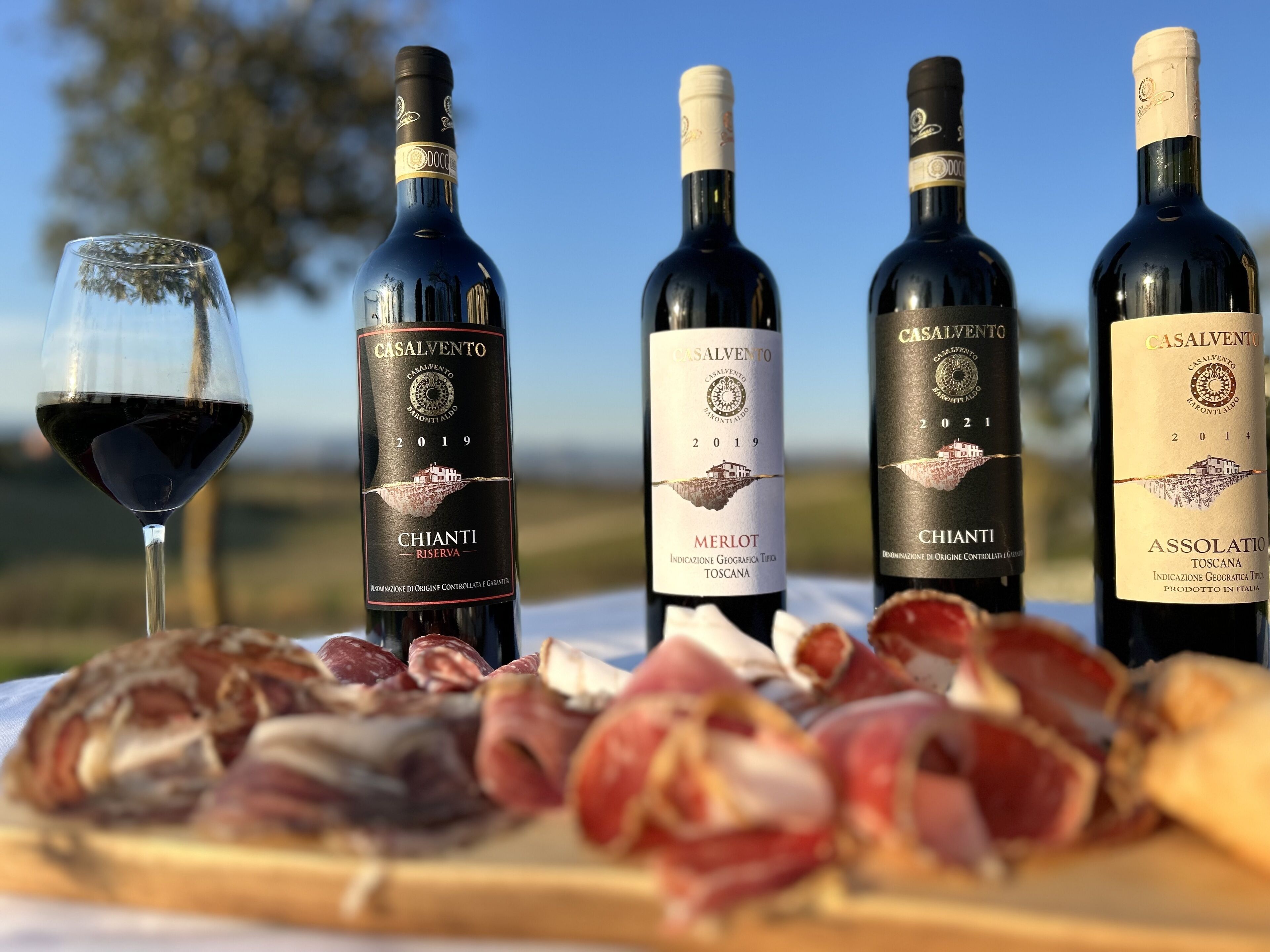 Four bottles of CASALVENTO wine and a glass of red wine on a table with various cured meats.