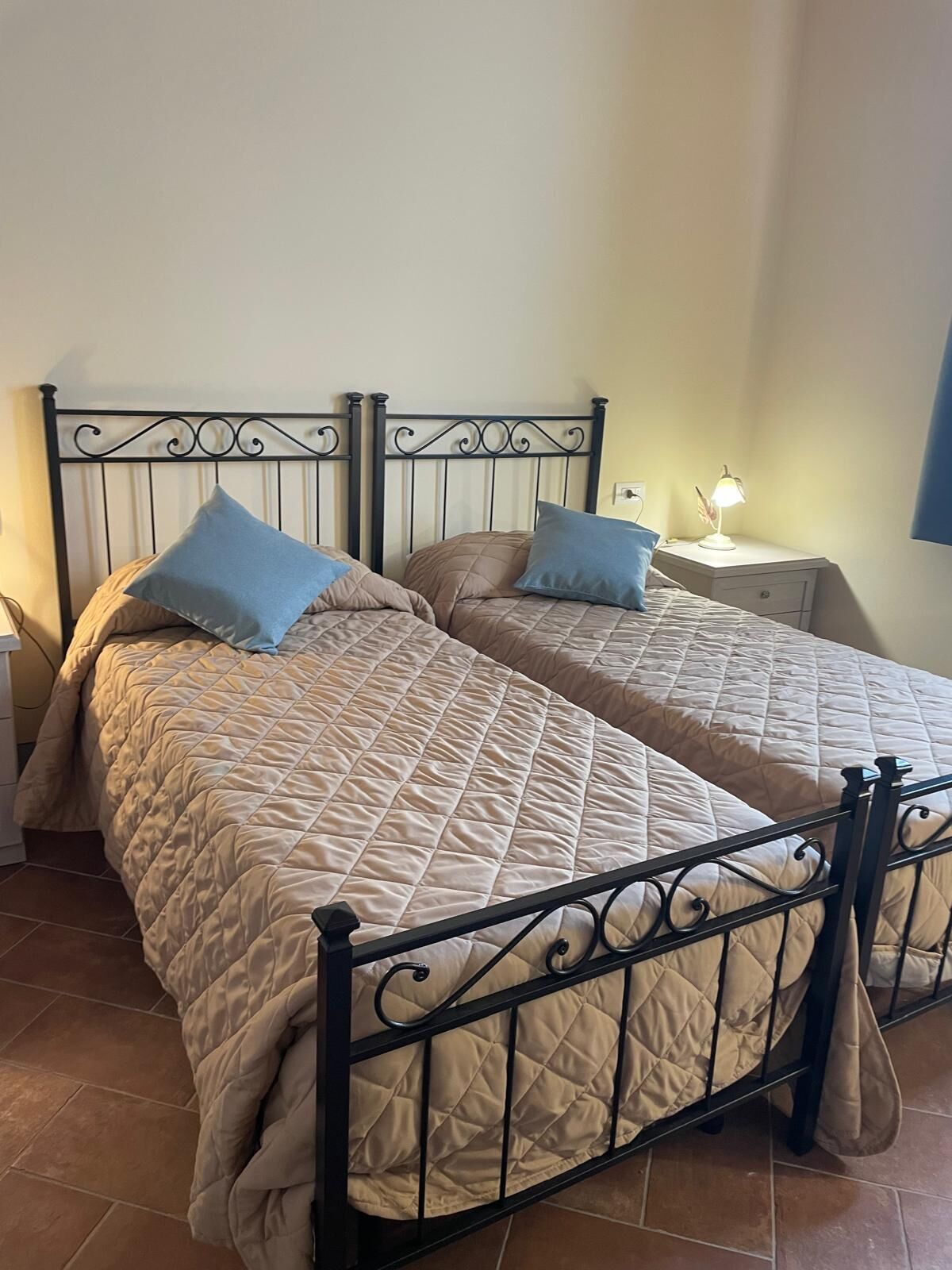 Een tweepersoonsbed met een gebogen beige sprei, twee blauwe kussens en een zwart metalen hoofdbord.