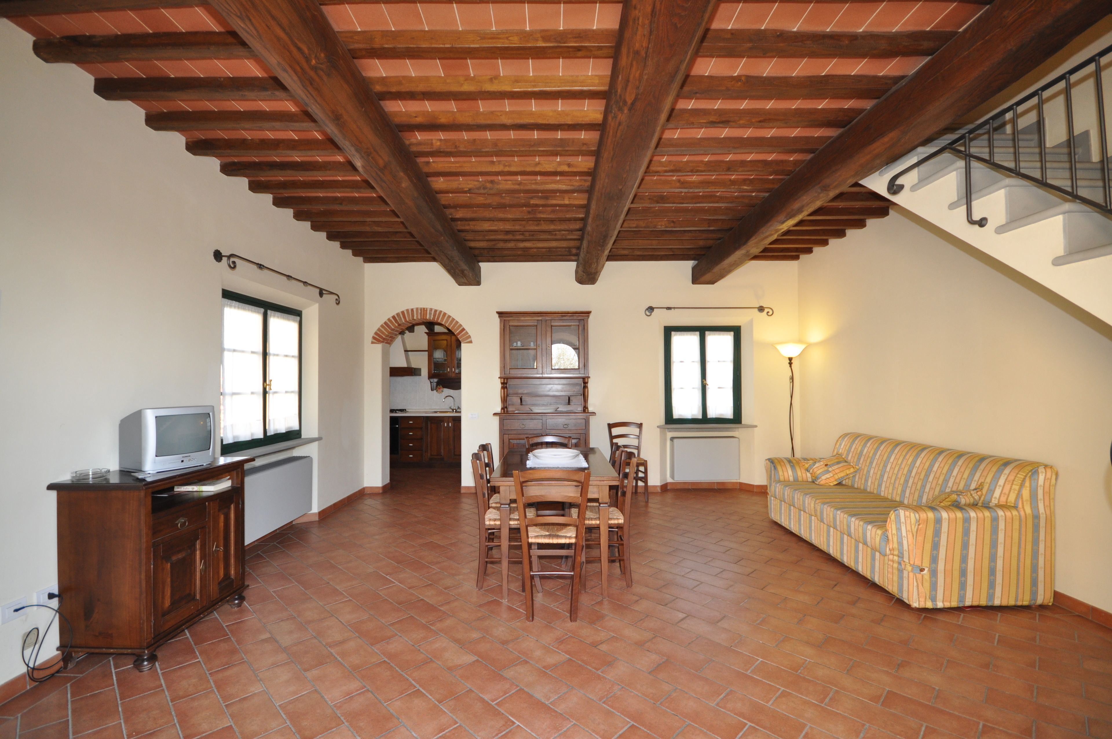 A spacious living area with a dining table and chairs, a sofa, a TV on a cabinet, and a staircase.
