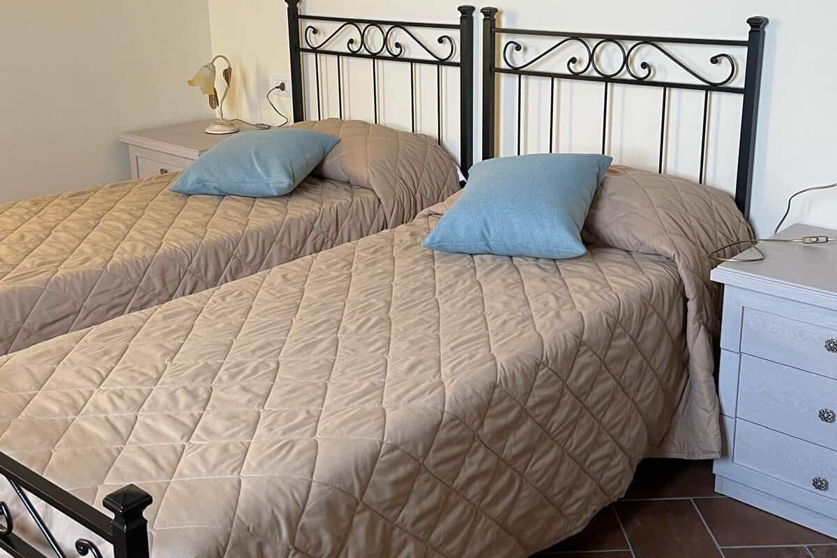 A double bed with a beige quilted bedspread and two pillows, a bedside table with a lamp, and a white dresser with drawers.