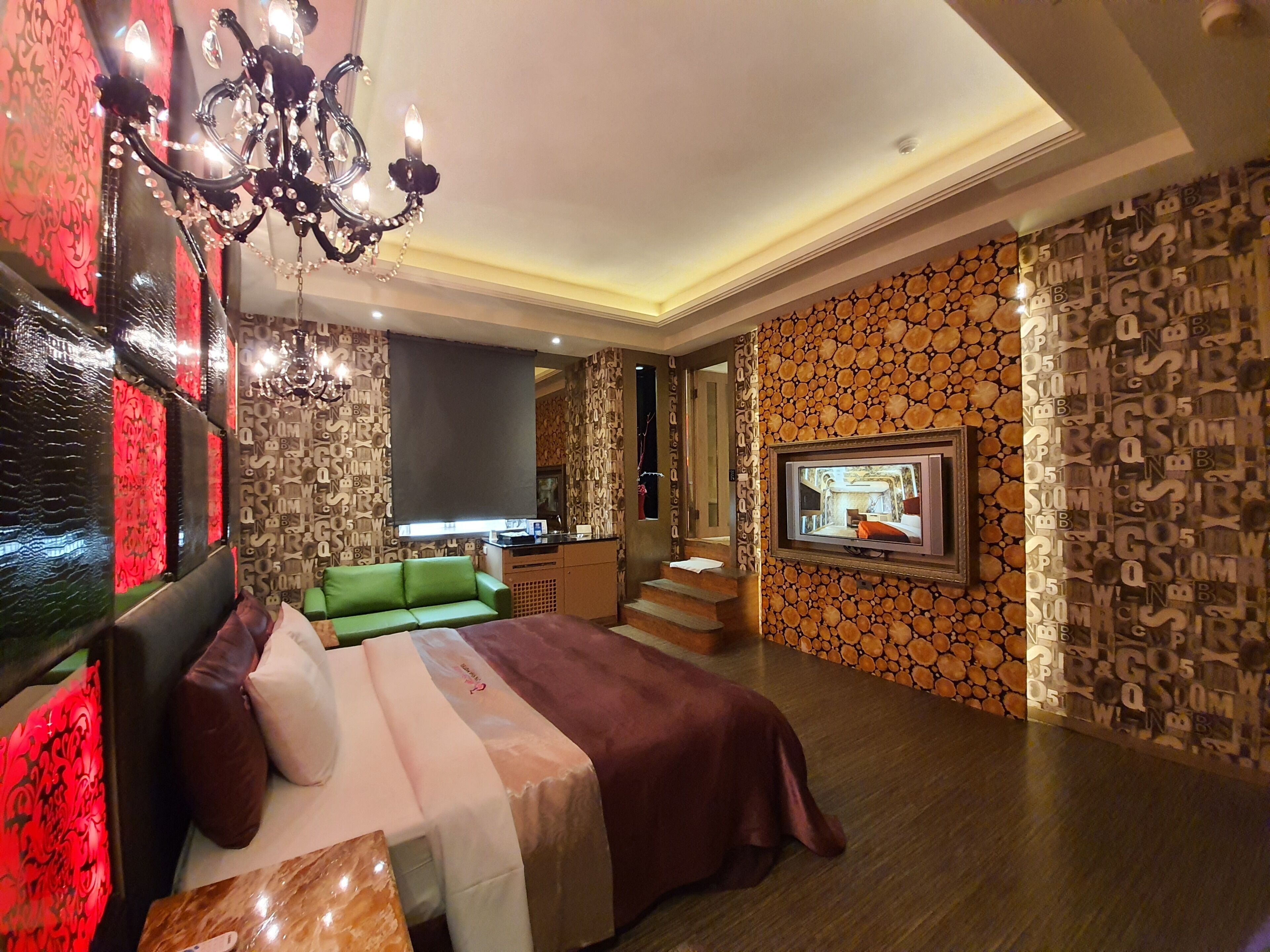 Quarto de hotel moderno com uma cama grande, um sofá verde, uma televisão e um candelabro.
