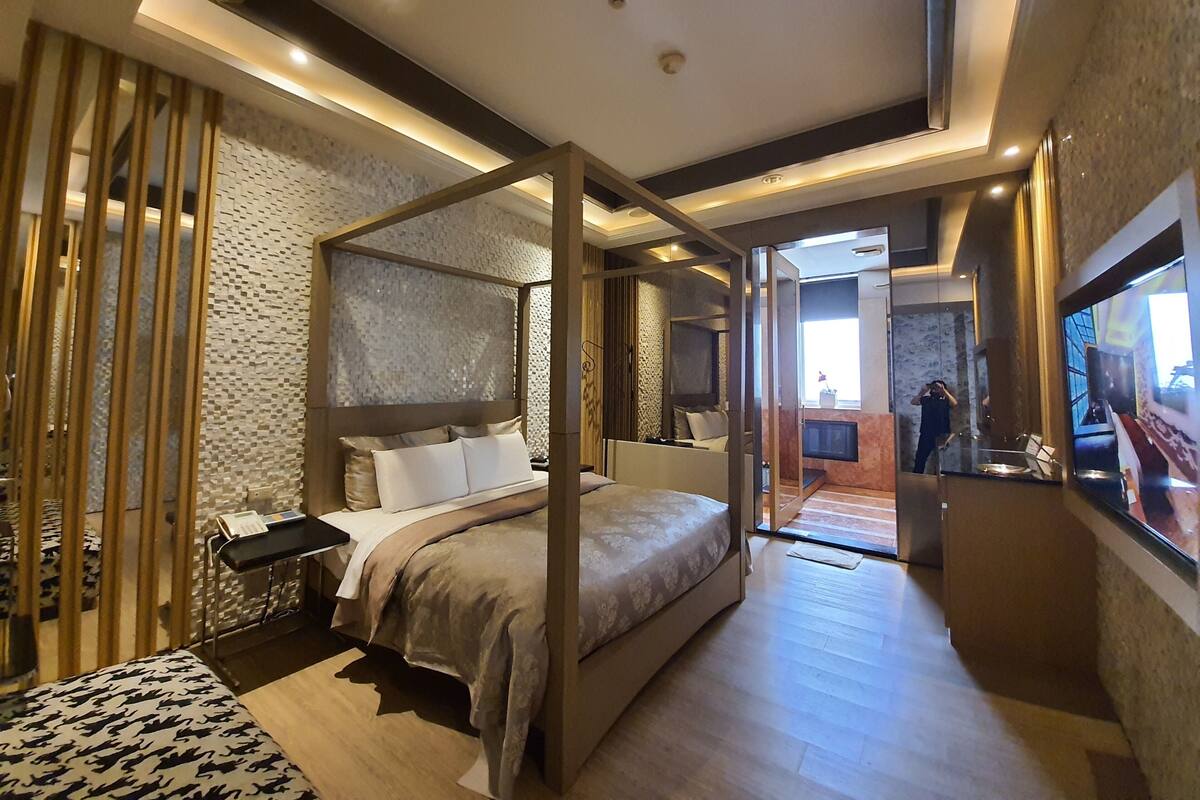 A modern hotel room with a four-poster bed, a large mirror, and a view of the city through the window.
