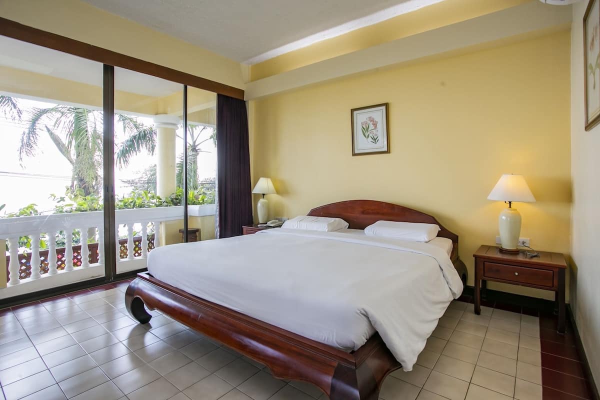 A hotel room with a large bed, a nightstand with a lamp, a framed picture on the wall, and a balcony with a view of palm trees.