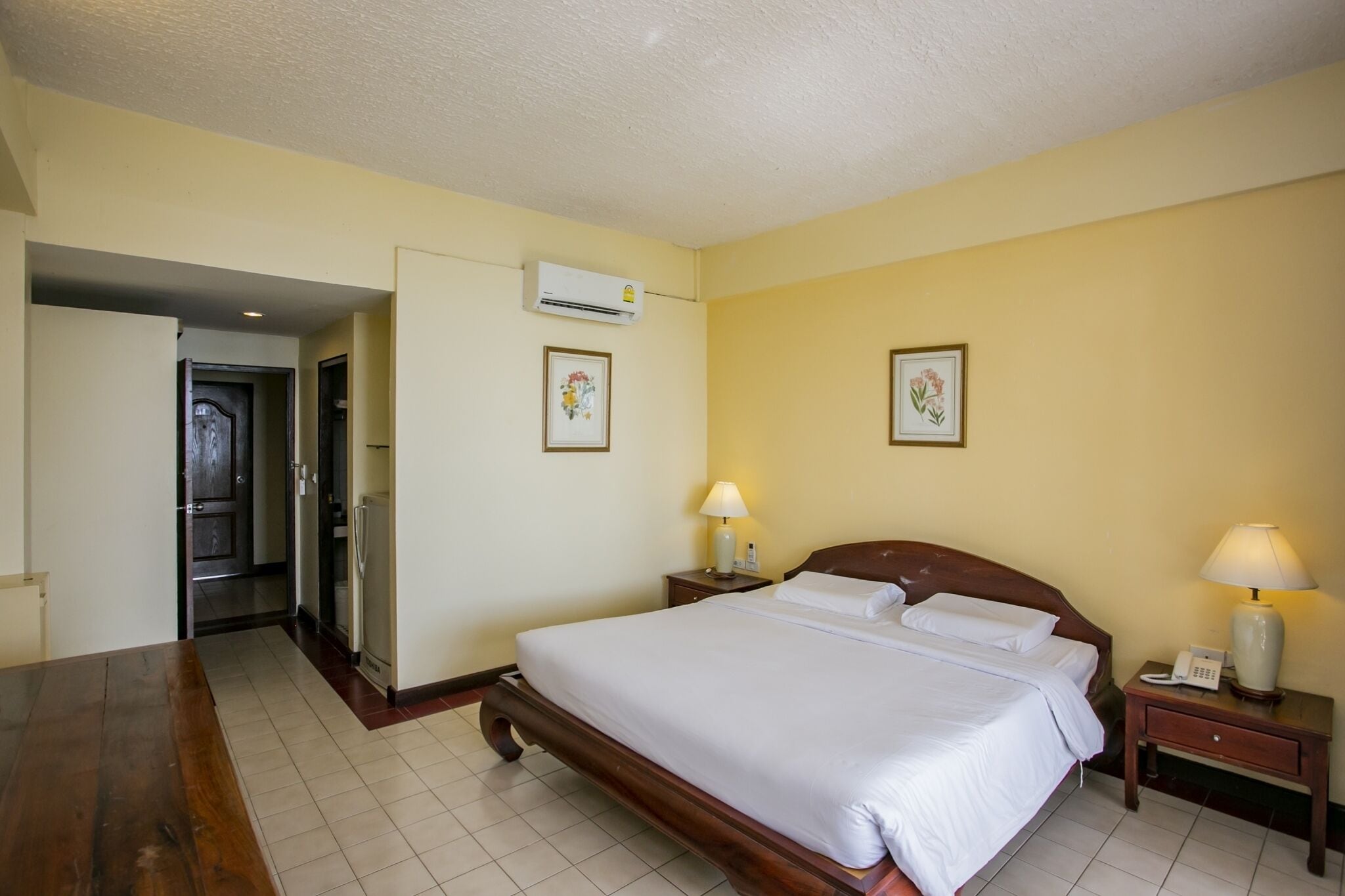 A hotel room with a bed, bedside tables, a desk, an air conditioning unit, and framed pictures on the walls.