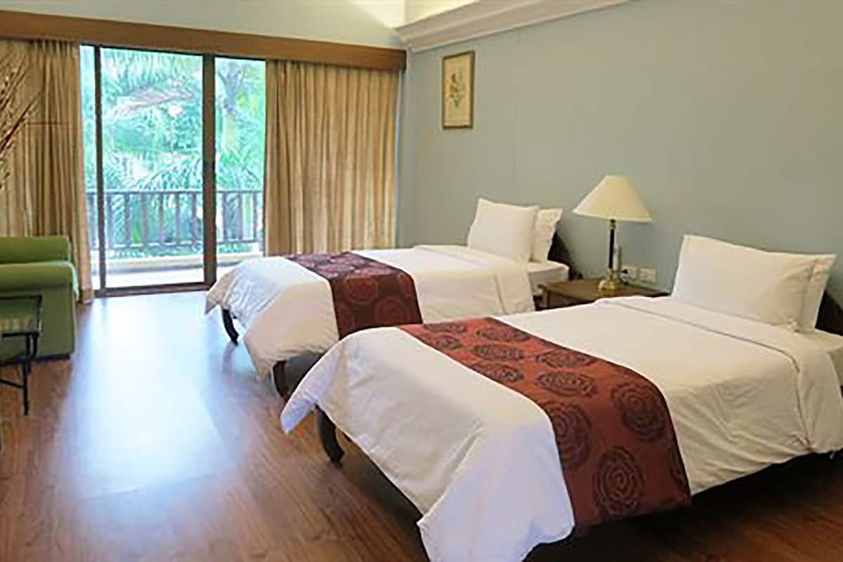 A hotel room with two beds, a green armchair, a balcony with curtains, and a painting on the wall.