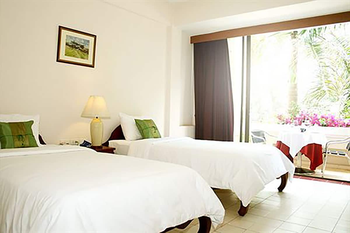 A hotel room with two beds, a nightstand, a lamp, a framed picture on the wall, and a view of a garden through a large window.