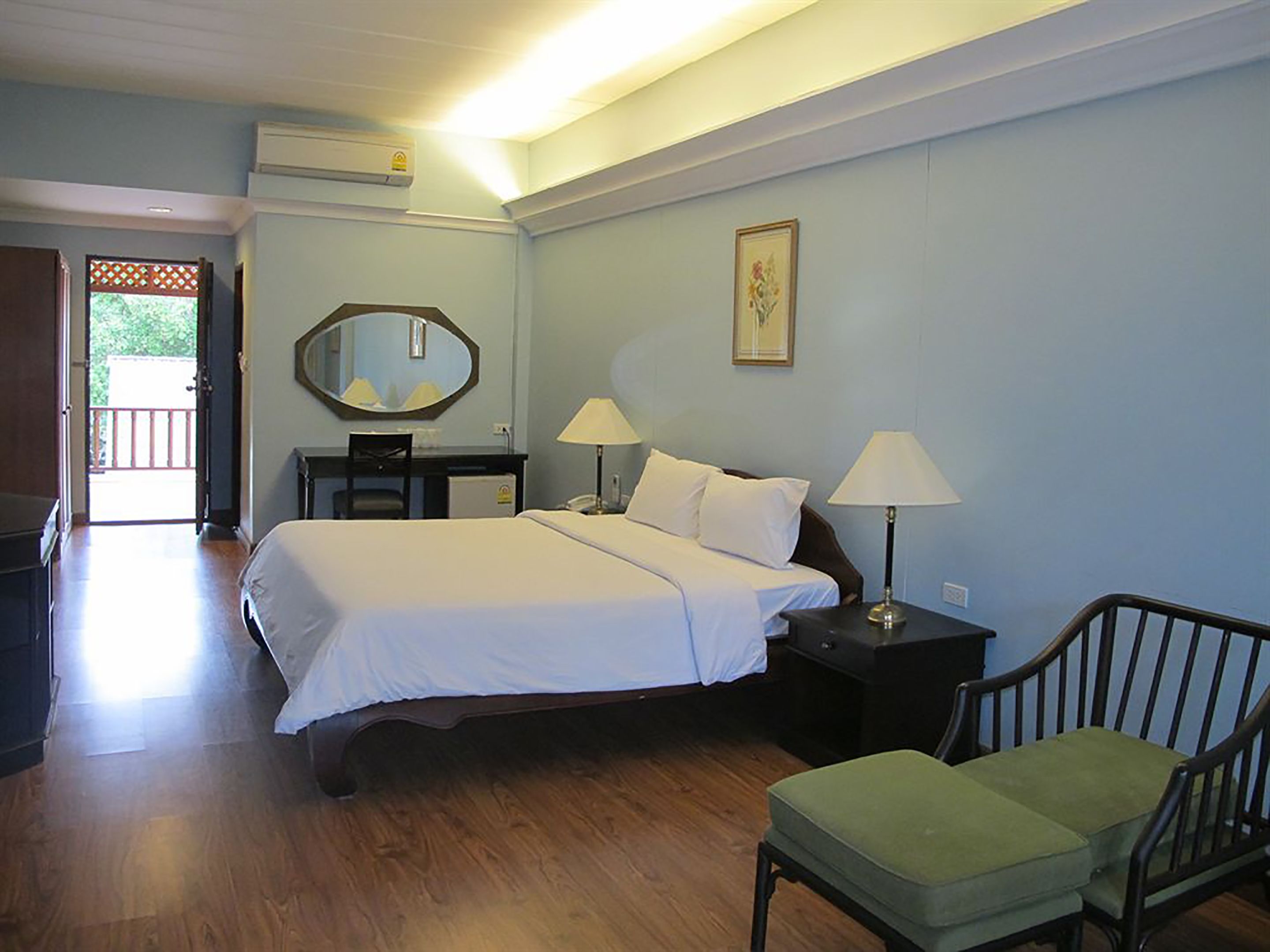Una habitación de hotel con una cama, dos mesitas de noche con lámparas, una silla, un escritorio con silla, un espejo e imagen en la pared.
