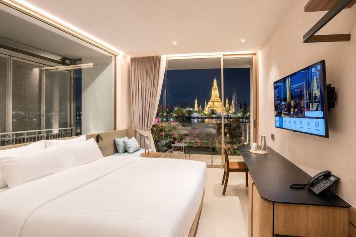 A hotel room with a large bed, a desk with a phone, a TV, and a balcony with a view of a temple at night.