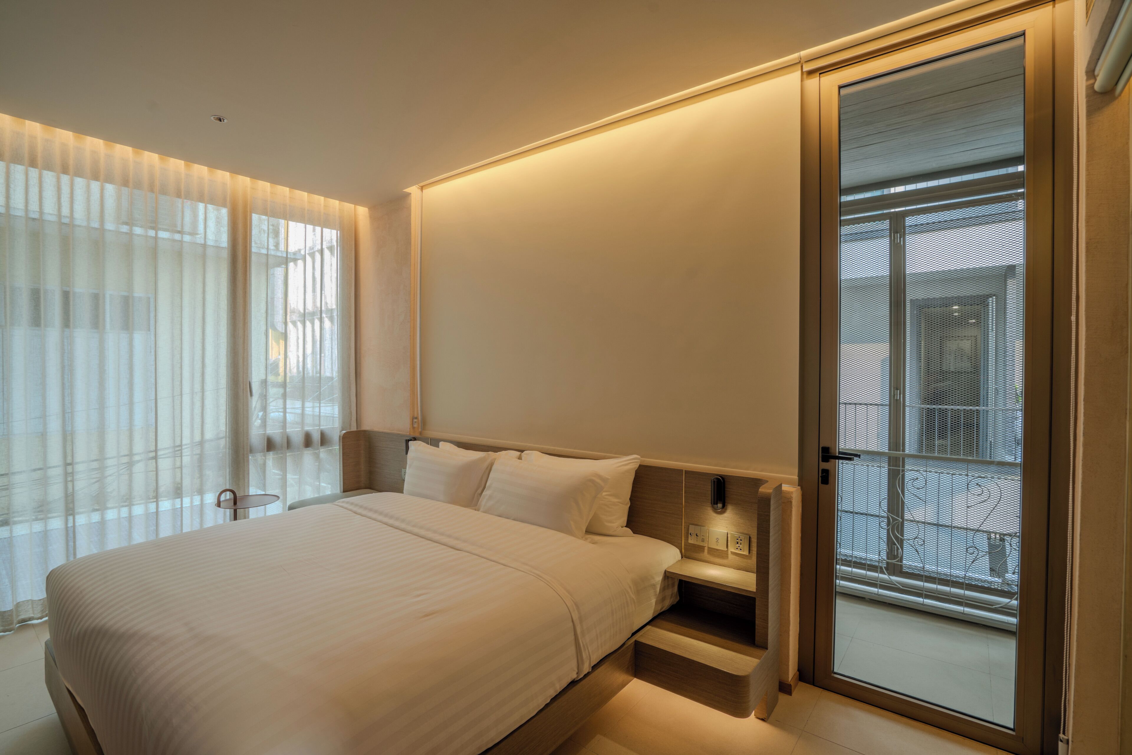 A modern hotel room with a large bed, bedside tables, a sliding glass door to the balcony, and sheer curtains on the windows.