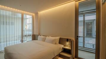 A modern hotel room with a large bed, bedside tables, a sliding glass door to the balcony, and sheer curtains on the windows.