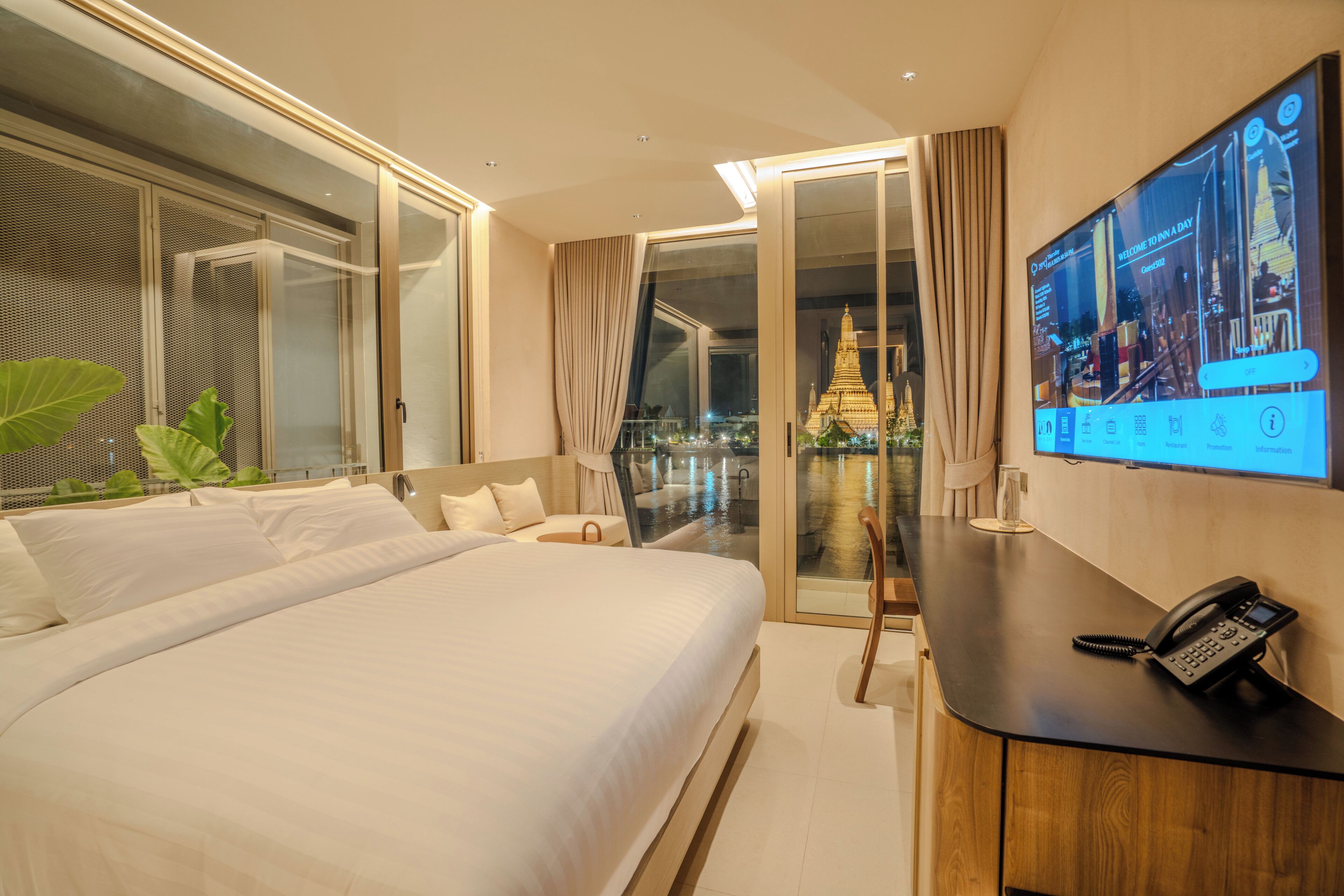 A hotel room with a large bed, a desk with a phone, a TV screen, and a view of a temple through a glass door.