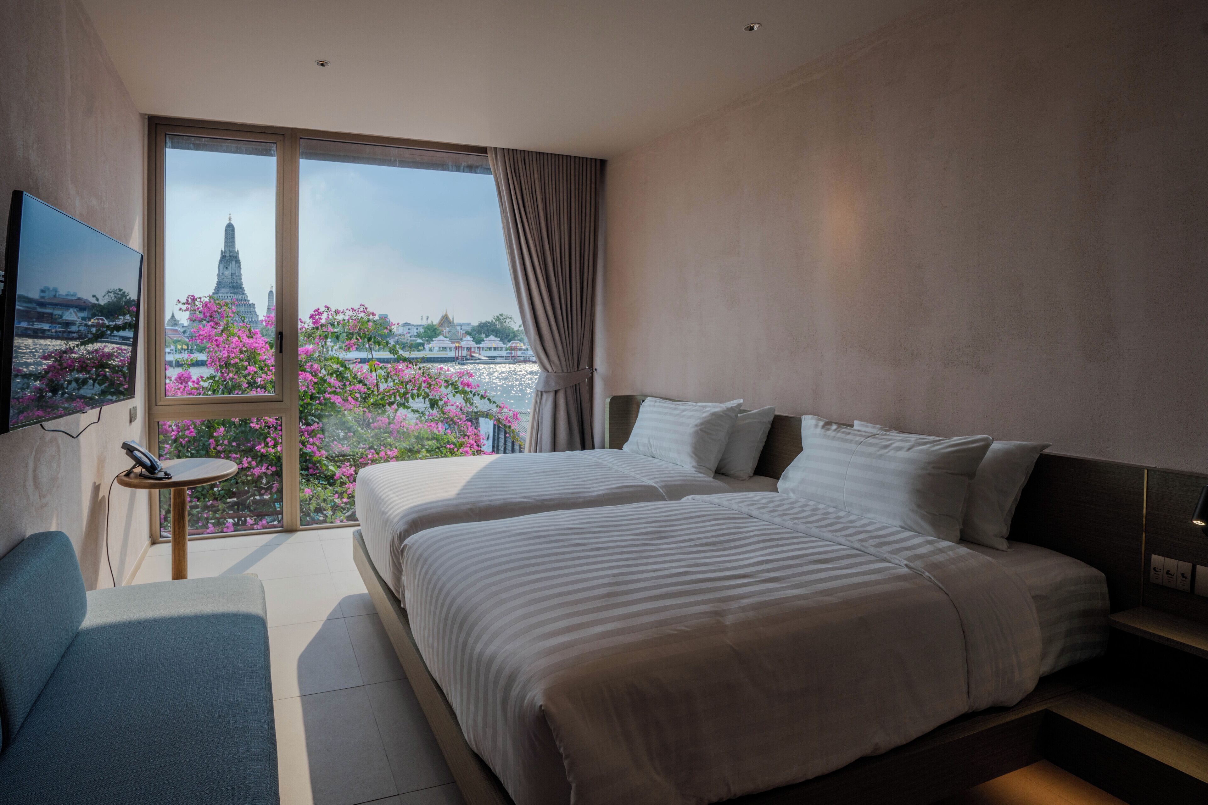 A modern hotel room with a large bed, a TV, a small table, and a view of a temple through a large window.