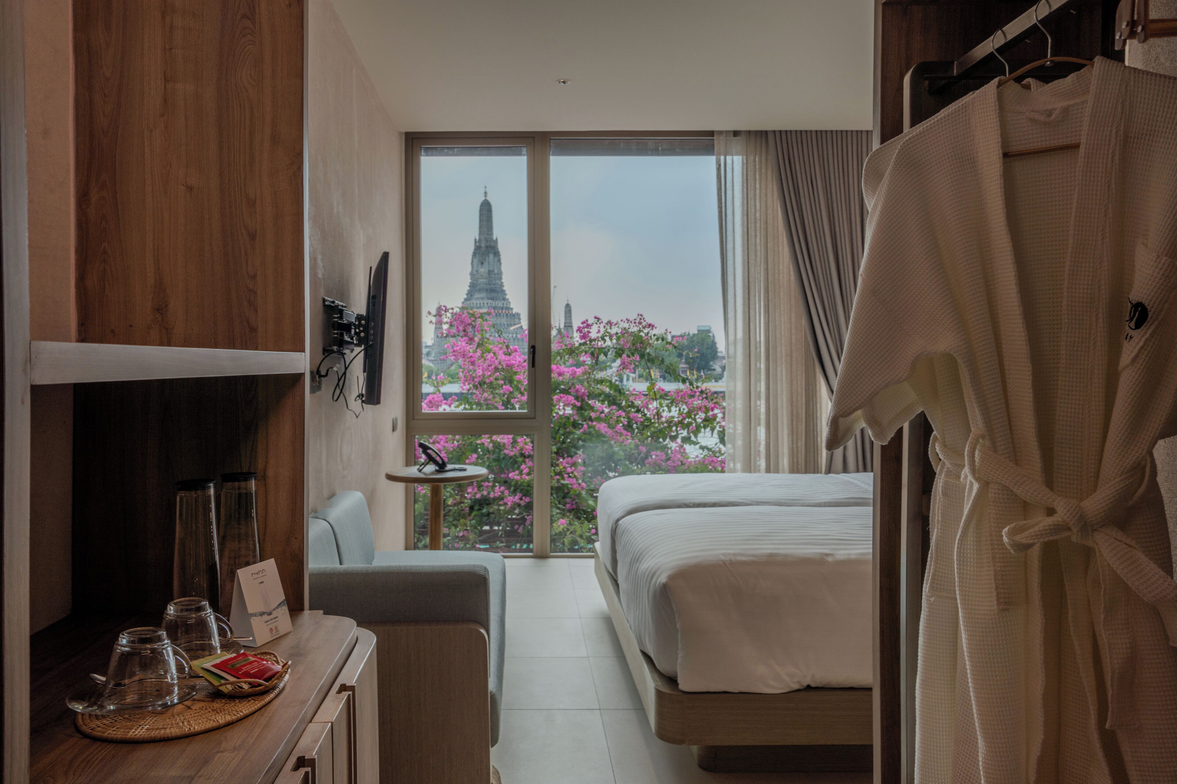 A hotel room with a bed, a desk with a chair, a robe hanging, and a window with a view of a temple and pink flowers.