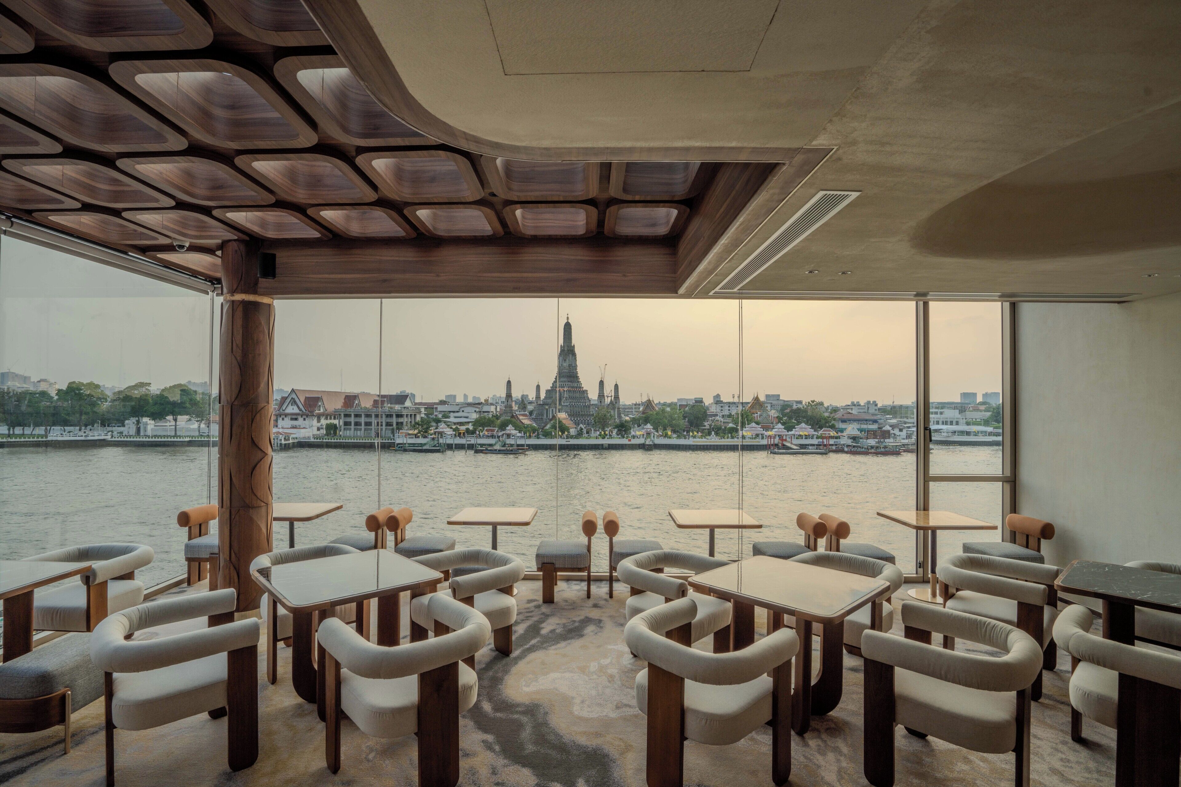 A restaurant with tables and chairs, a view of a river and city skyline, and a modern ceiling design.