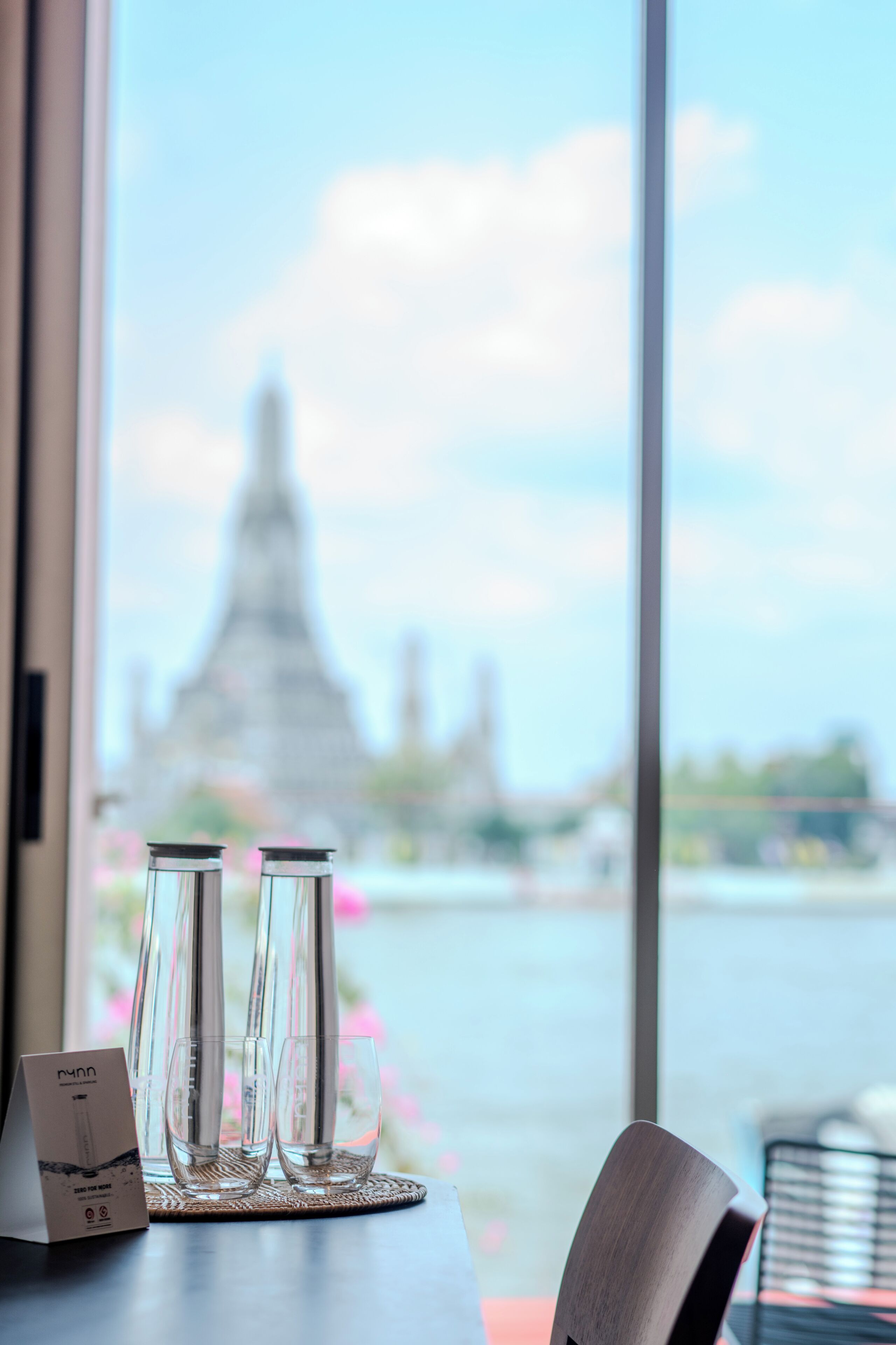 A table with two glasses and a menu, a chair, and a view of a temple through a window.