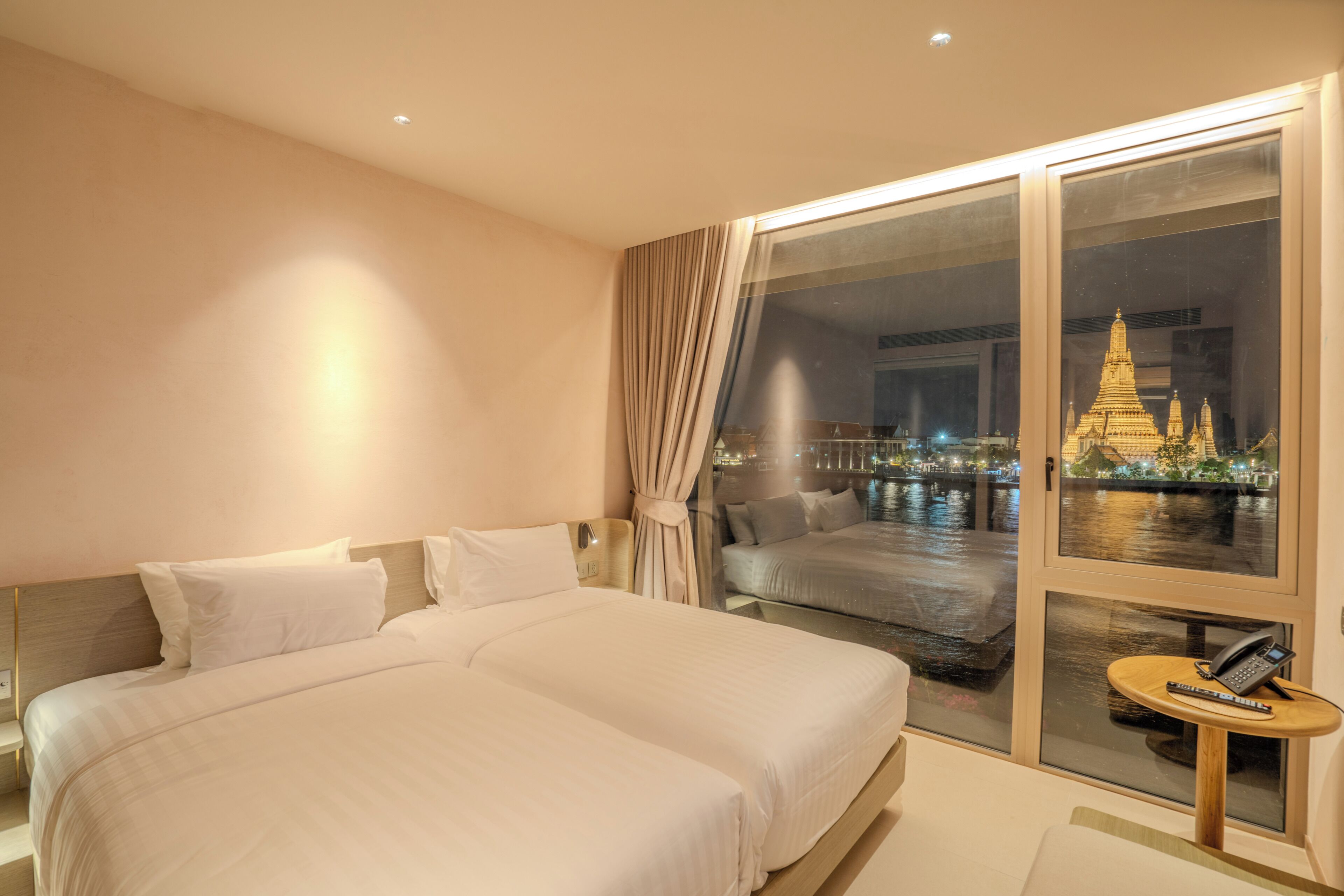 A hotel room with a large bed, a small table with a phone, and a view of a temple through a window.