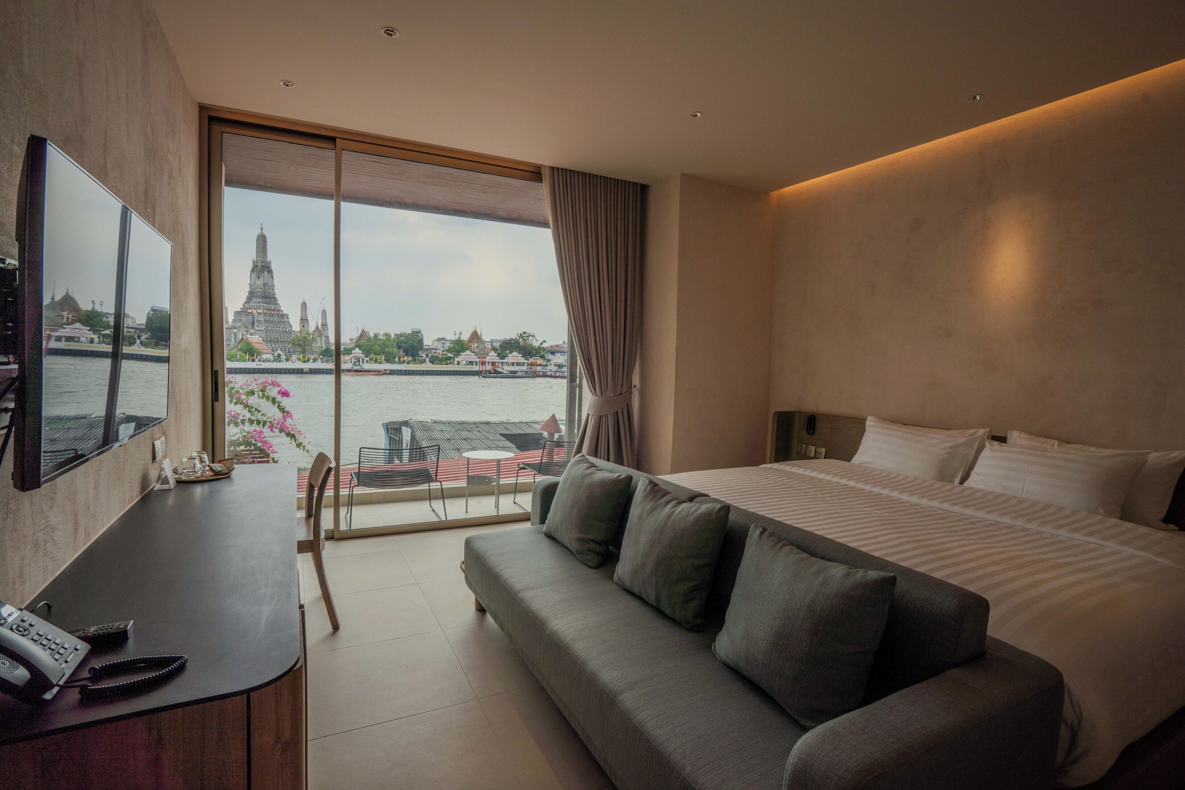 Habitación de hotel moderna con una cama grande, un sofá, un escritorio con un teléfono y documentos, y un ventanal con vistas a un templo y al agua.