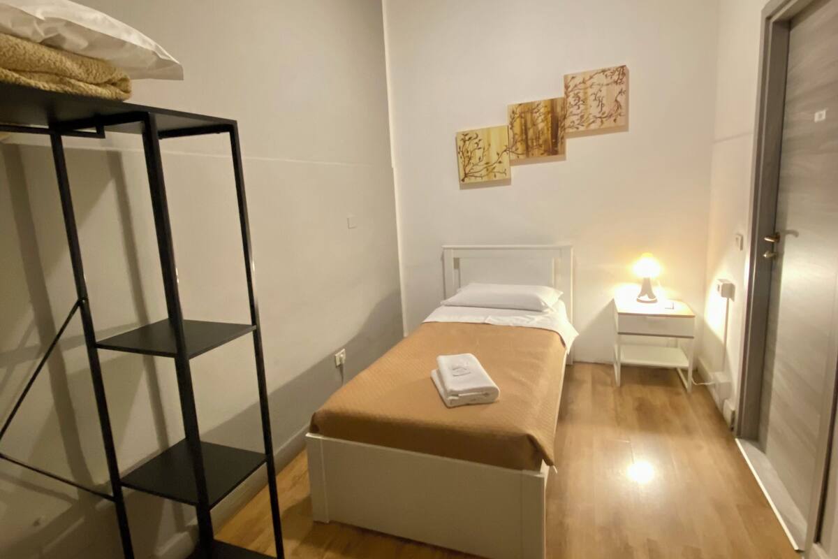 A small, well-lit bedroom with a single bed, a bedside table with a lamp, and a shelf unit.
