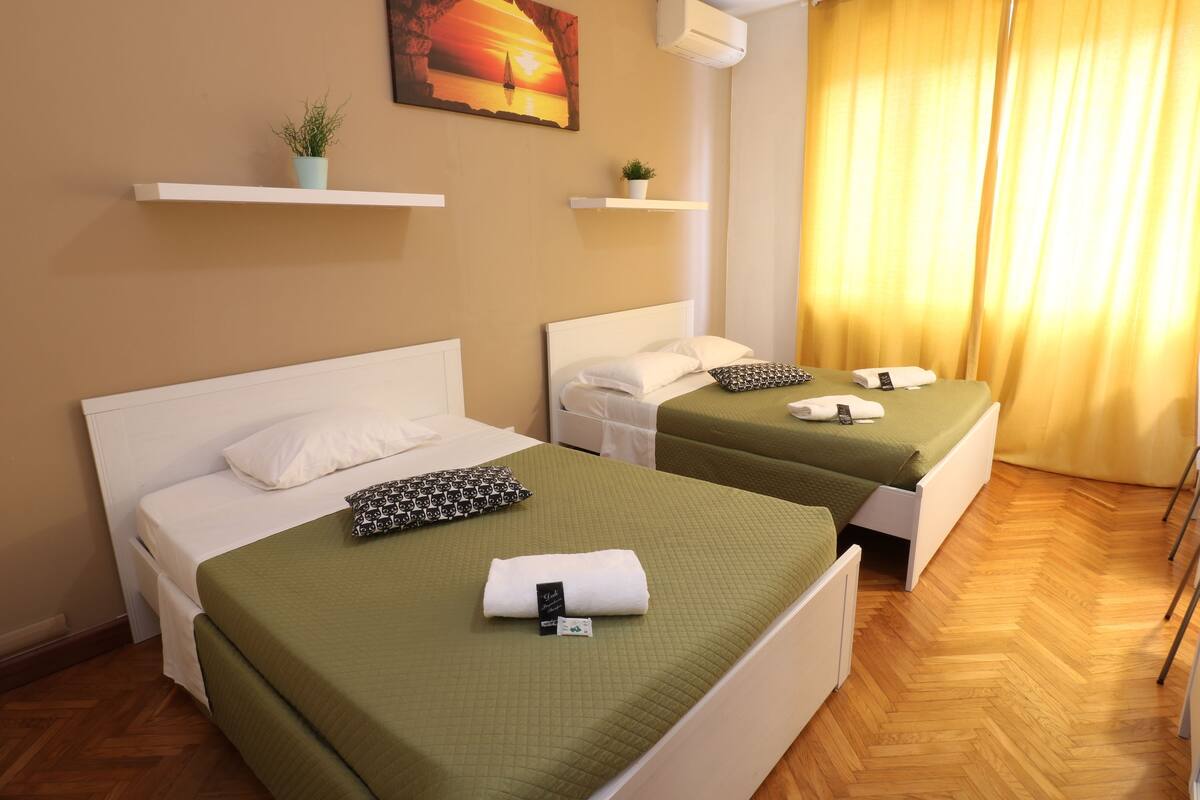 A hotel room with two beds, each with a green bedspread, white pillows, and a small potted plant on a shelf above.
