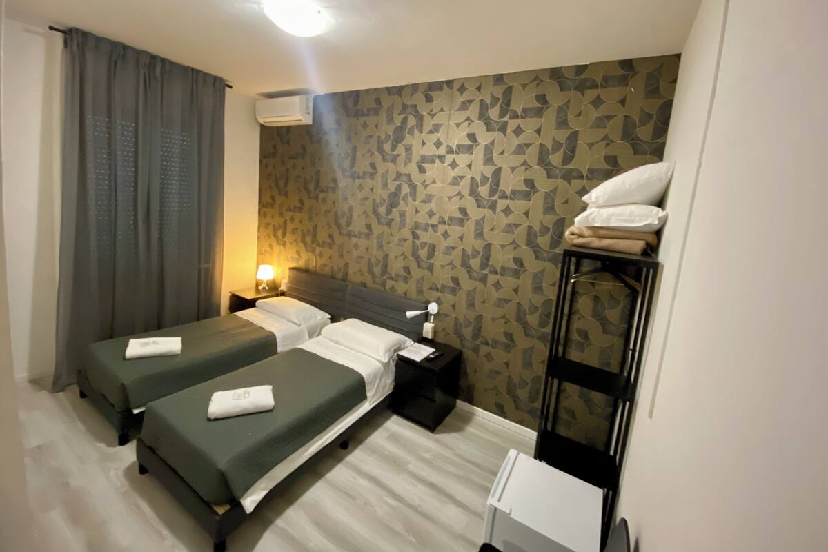 A hotel room with two beds, a bedside table with a lamp, a wall-mounted air conditioner, and a small white cabinet.