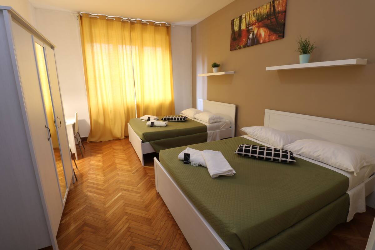 A hotel room with two beds, a wooden floor, a window with yellow curtains, a painting on the wall, and a small plant on a shelf.
