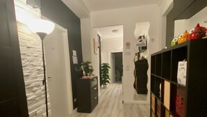A hallway with a light-colored wooden floor, a black cabinet with storage, a potted plant, and a wall-mounted shelf with items. - Guest House Pirelli (Milan)