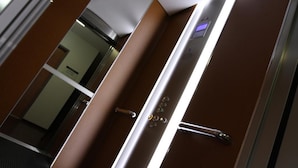 Interior de elevador com corrimão metálico, painel de comandos com botões e porta de vidro.