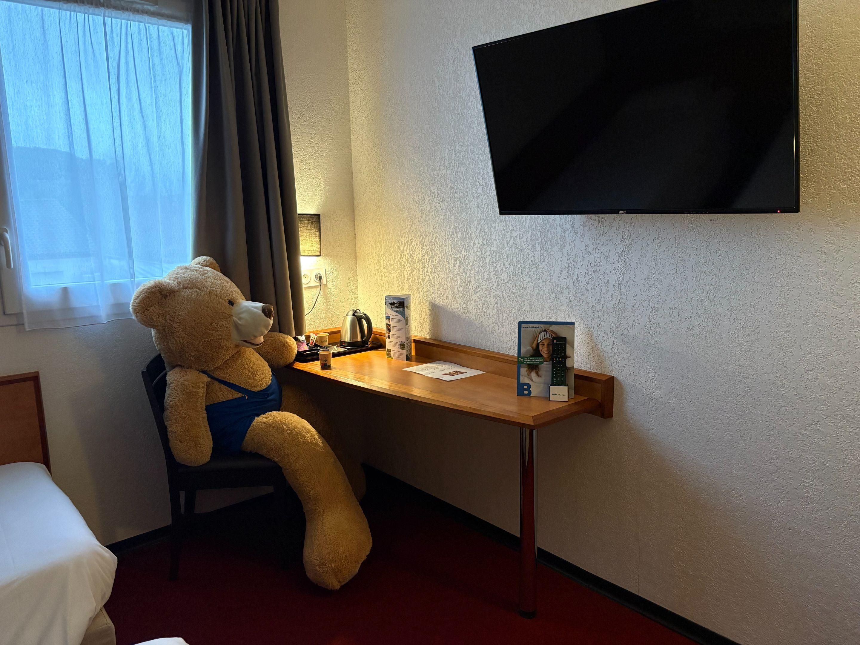 Une chambre d’hôtel avec un lit, un bureau sur lequel est posé un ours en peluche, une bouilloire, une télécommande et un téléviseur fixé au mur.