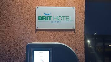 A Brit Hotel Comfort sign on a textured wall, a digital display with a name and room type, and a door to the right.
