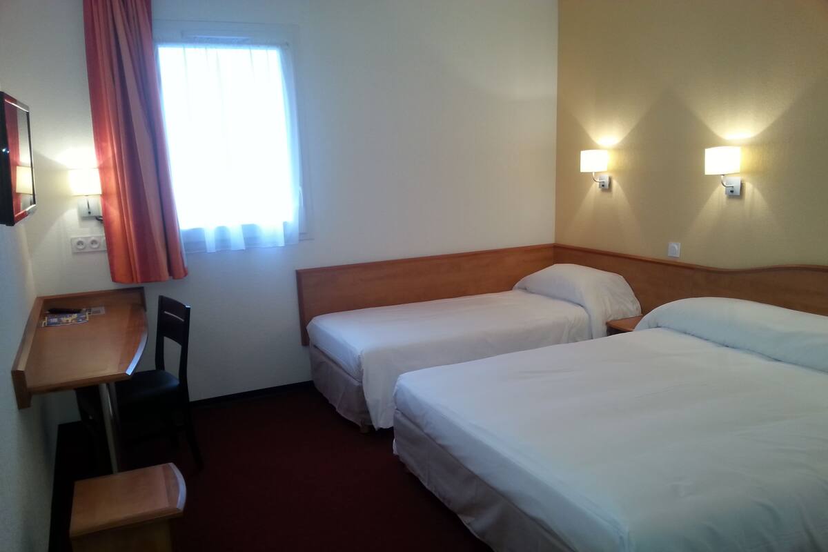 Ein Hotelzimmer mit zwei Einzelbetten, einem Schreibtisch mit Stuhl, einem kleinen Tisch und einem Fenster mit Vorhängen.