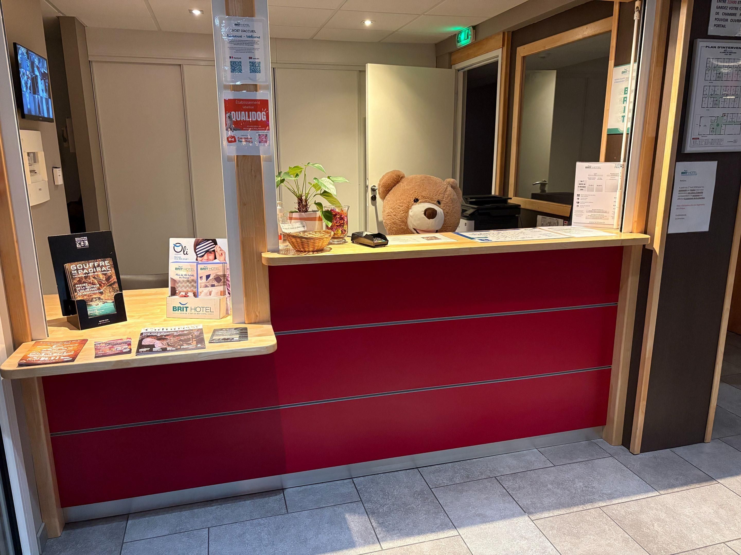 Un comptoir d’accueil avec un plan de travail rouge, un grand ours en peluche et des affiches d’information.