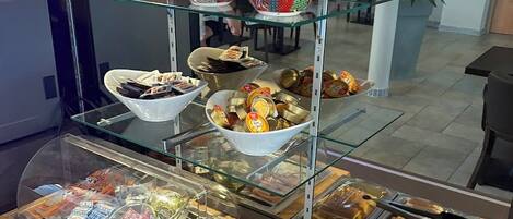 Zona de bufé con diversas opciones de comida expuestas en recipientes de vidrio, como pan, pasteles y salsas.