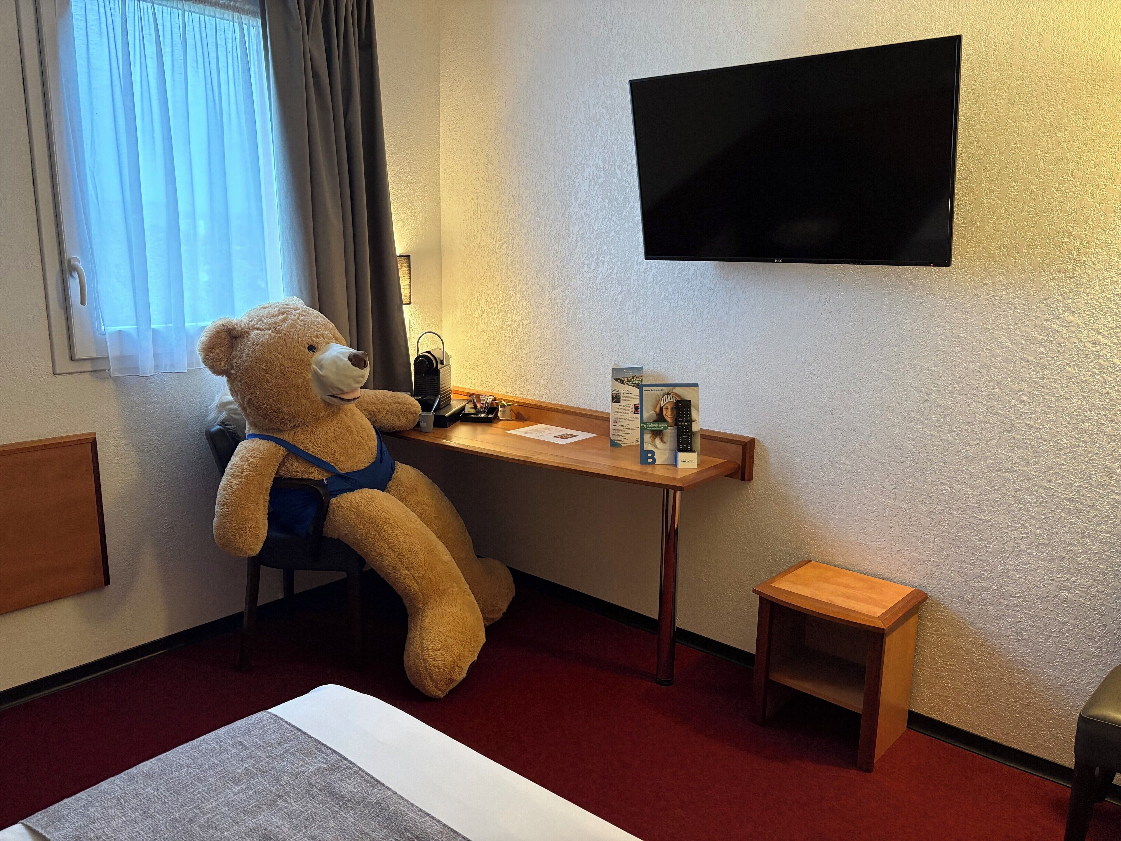 Una camera d'albergo con un letto, una scrivania con un orsacchiotto, una televisione e uno sgabello di legno.