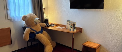 A hotel room with a bed, a desk with a teddy bear, a TV, and a small wooden stool.