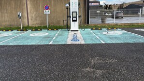 Uma área de estacionamento com estações de carregamento para veículos elétricos, um aviso de proibido estacionar e um edifício ao fundo.
