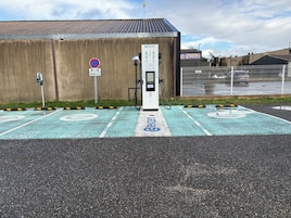 Uma área de estacionamento com estações de carregamento para veículos elétricos, um aviso de proibido estacionar e um edifício ao fundo.