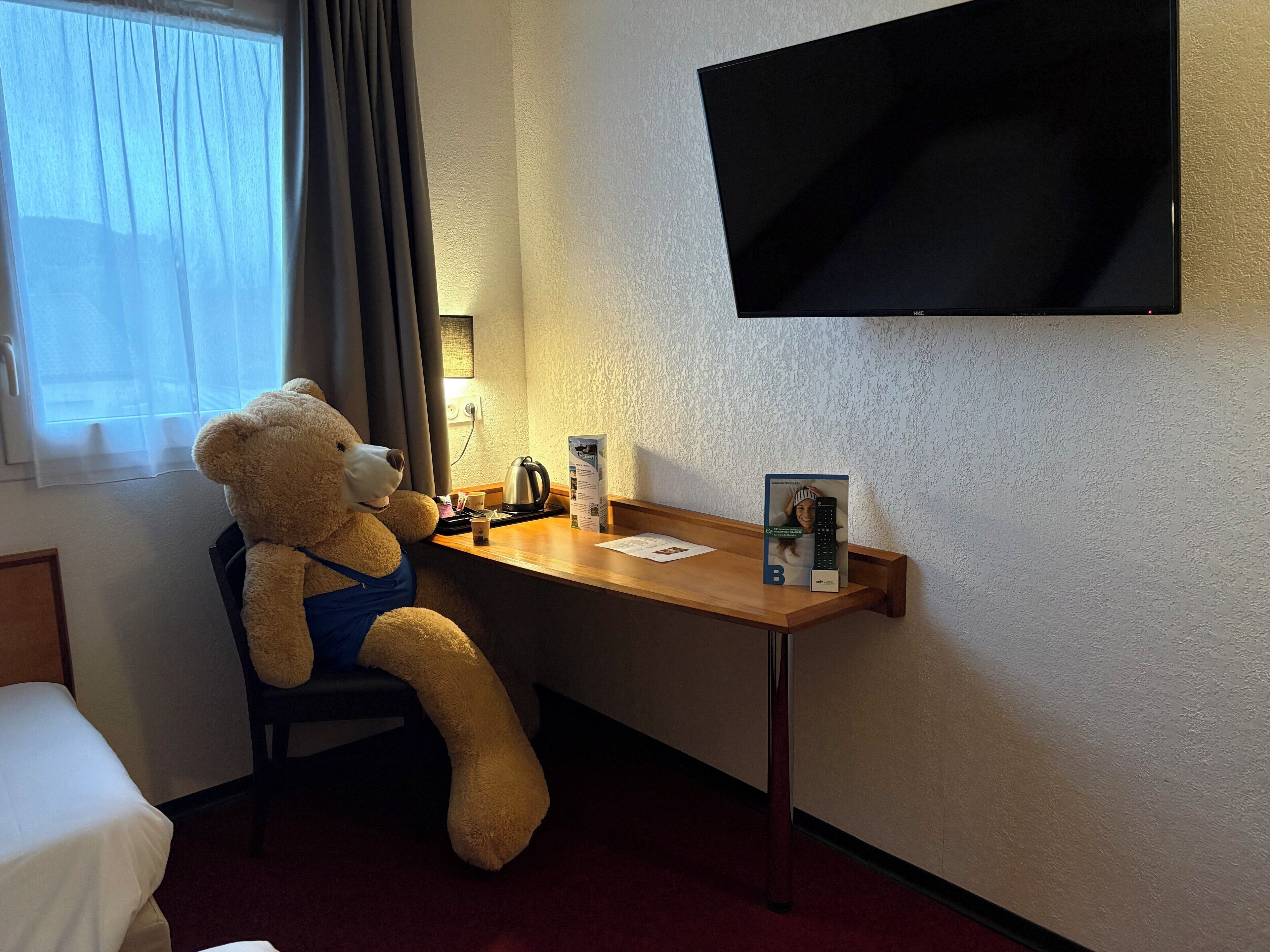 Una habitación de hotel con una cama, un escritorio con un osito de peluche, una tetera, un control remoto y un televisor montado en la pared.