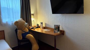 Una habitación de hotel con una cama, un escritorio con un osito de peluche, una tetera, un control remoto y un televisor montado en la pared.