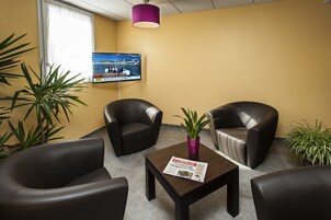 Uma área de espera com poltronas de couro, uma mesa com jornal e uma televisão na parede.