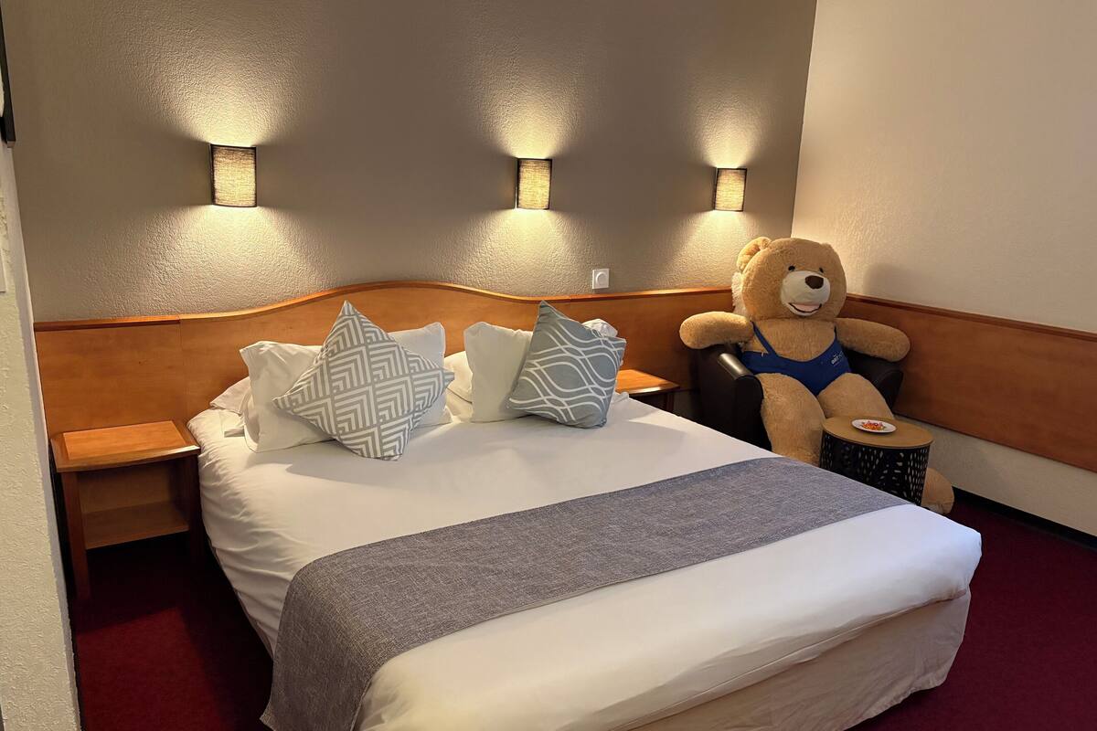 Ein ordentlich gemachtes Bett mit einer grauen Decke, zwei gemusterten Kissen und einem Teddybären auf einem kleinen runden Tisch daneben.